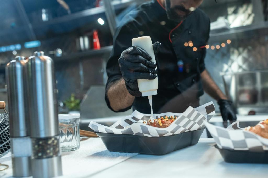 Chef in a kitchen preparing gourmet hot dogs with sauce on a black tray.