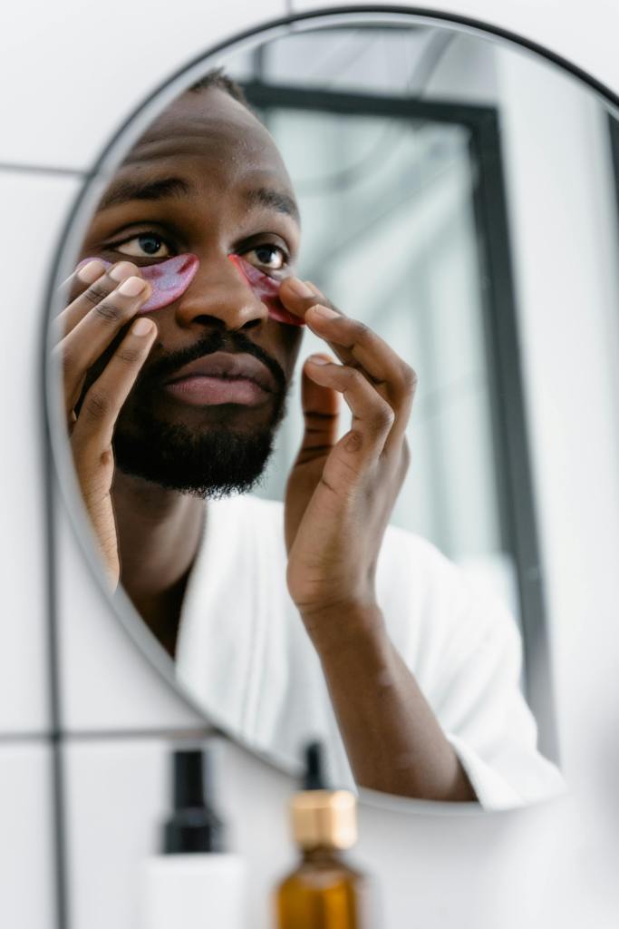 A man applies under eye masks while looking in the mirror, practicing skincare.